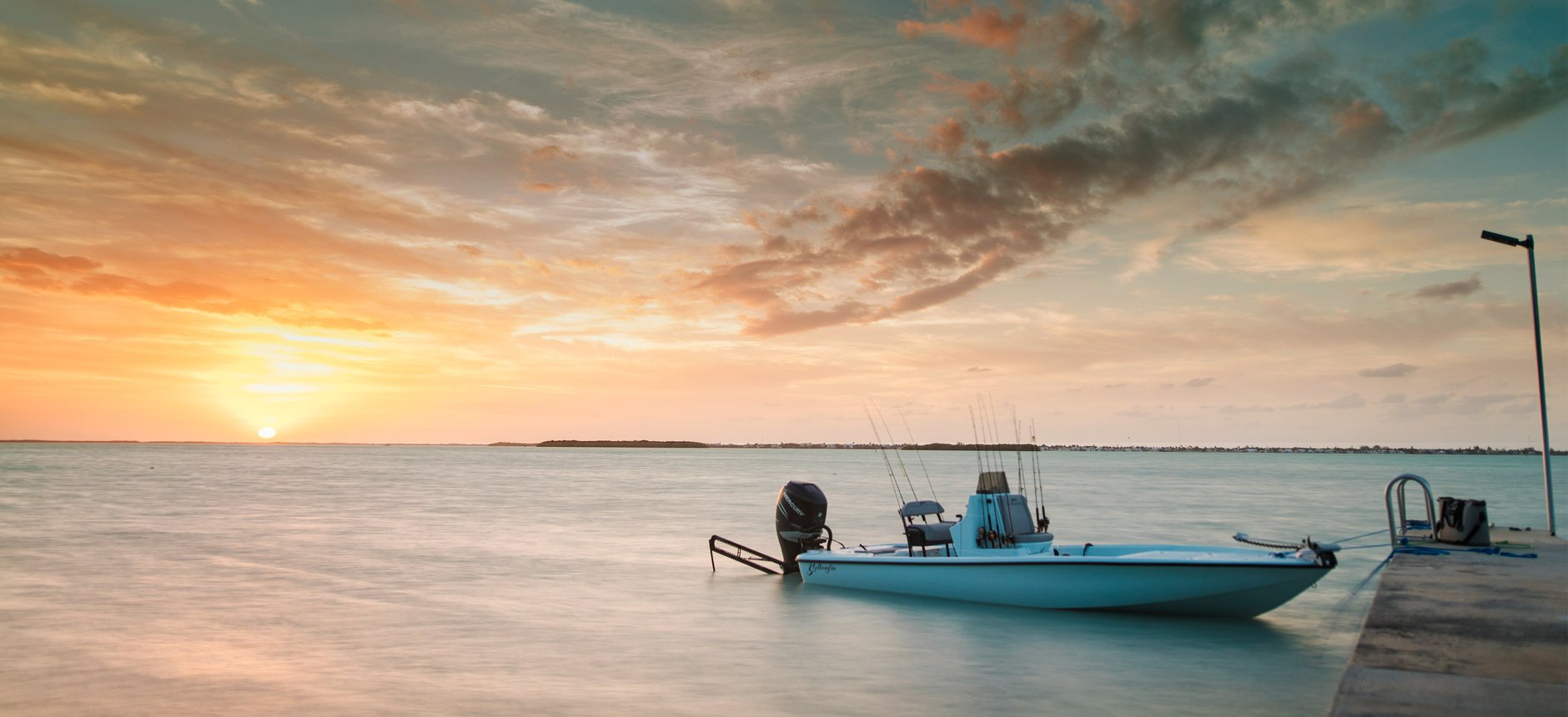 A fishing boat with an outboard motor, equipped with fishing rods, is moored at a pier during sunset.